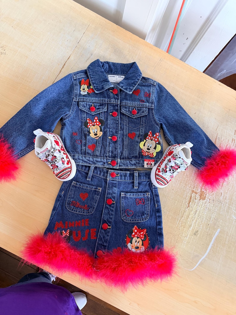 May include: A denim jacket and skirt set with red accents and Minnie Mouse appliqu&eacute;s. The jacket has red buttons and faux fur trim on the sleeves. The skirt has red buttons and a red faux fur trim. Matching sneakers complete the outfit.