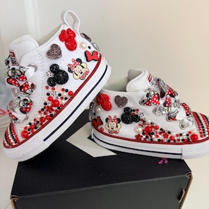 May include: White high-top sneakers with red and black accents, embellished with Minnie Mouse graphics, red and black heart-shaped gems, and pearls. The shoes have red and white striped detailing on the toe.