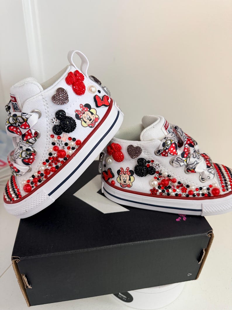 May include: White high-top sneakers with red and black accents, embellished with Minnie Mouse graphics, red and black heart-shaped gems, and pearls. The shoes have red and white striped detailing on the toe.