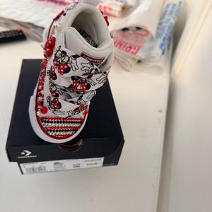 May include: White Converse toddler shoe, embellished with red and clear rhinestones, red beads, and pearl accents. The shoe features a Minnie Mouse print on the straps and is displayed on a black Converse box.
