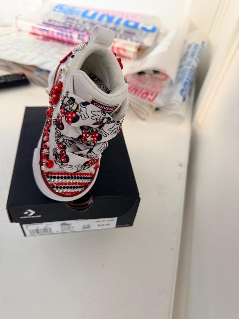 May include: White Converse toddler shoe, embellished with red and clear rhinestones, red beads, and pearl accents. The shoe features a Minnie Mouse print on the straps and is displayed on a black Converse box.