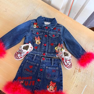 May include: A denim jacket, skirt, and shoes set. The blue denim jacket and skirt feature red heart and Minnie Mouse patches. The skirt has "MINNIE MUJE" embroidered in red. The jacket has red feather trim on the sleeves, and the skirt has red feather trim.