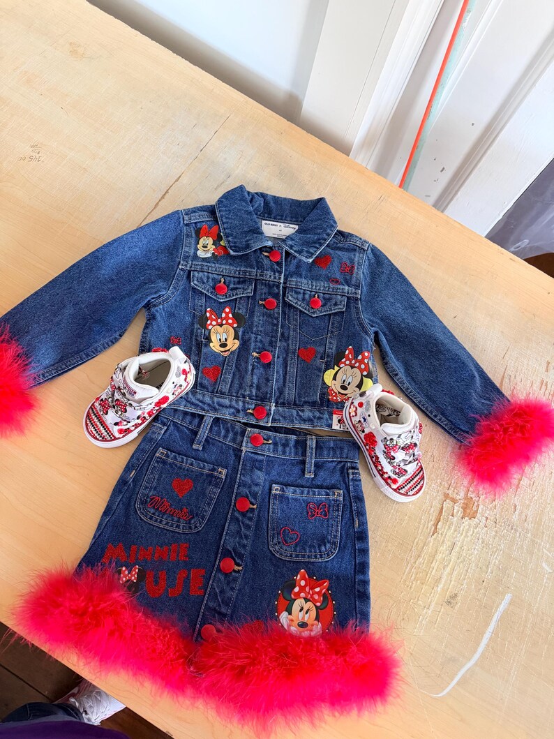 May include: A denim jacket, skirt, and shoes set. The blue denim jacket and skirt feature red heart and Minnie Mouse patches. The skirt has "MINNIE MUJE" embroidered in red. The jacket has red feather trim on the sleeves, and the skirt has red feather trim.
