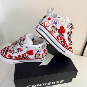 May include: White Converse high-top sneakers, embellished with red, silver, and pearl accents. The shoes feature Minnie Mouse graphics and red and black beadwork. The sneakers are displayed on a black Converse box.