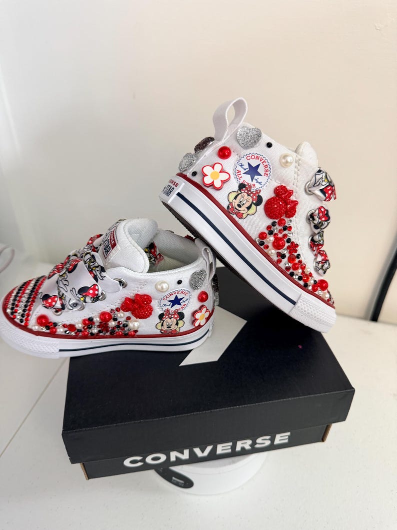 May include: White Converse high-top sneakers, embellished with red, silver, and pearl accents. The shoes feature Minnie Mouse graphics and red and black beadwork. The sneakers are displayed on a black Converse box.
