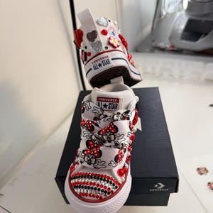May include: White Converse All Star sneakers, customized with Minnie Mouse ribbon laces, red and black rhinestones, and decorative embellishments. The shoes feature the Converse All Star logo on the tongue and heel.