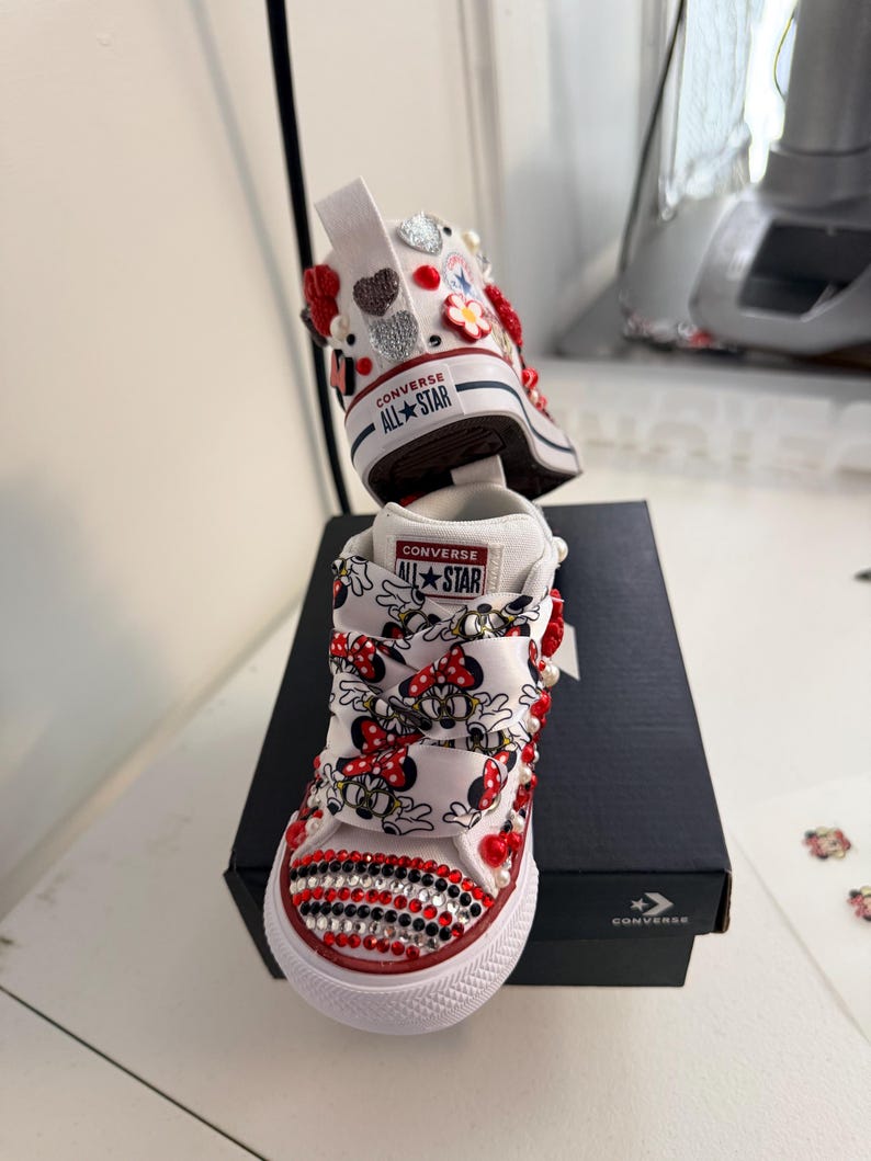 May include: White Converse All Star sneakers, customized with Minnie Mouse ribbon laces, red and black rhinestones, and decorative embellishments. The shoes feature the Converse All Star logo on the tongue and heel.