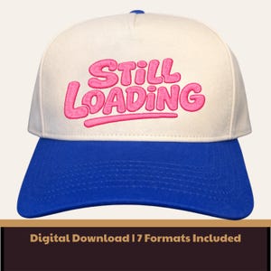 May include: A baseball cap with a white crown and a blue brim. The phrase "STILL LOADING" is embroidered in pink across the front. The cap has a blue button on top.