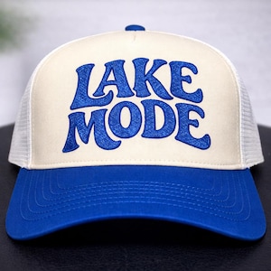 May include: A blue and cream-colored trucker hat with the words "LAKE MODE" embroidered in blue. The hat features a blue brim and top button, a cream-colored front panel, and a white mesh back.