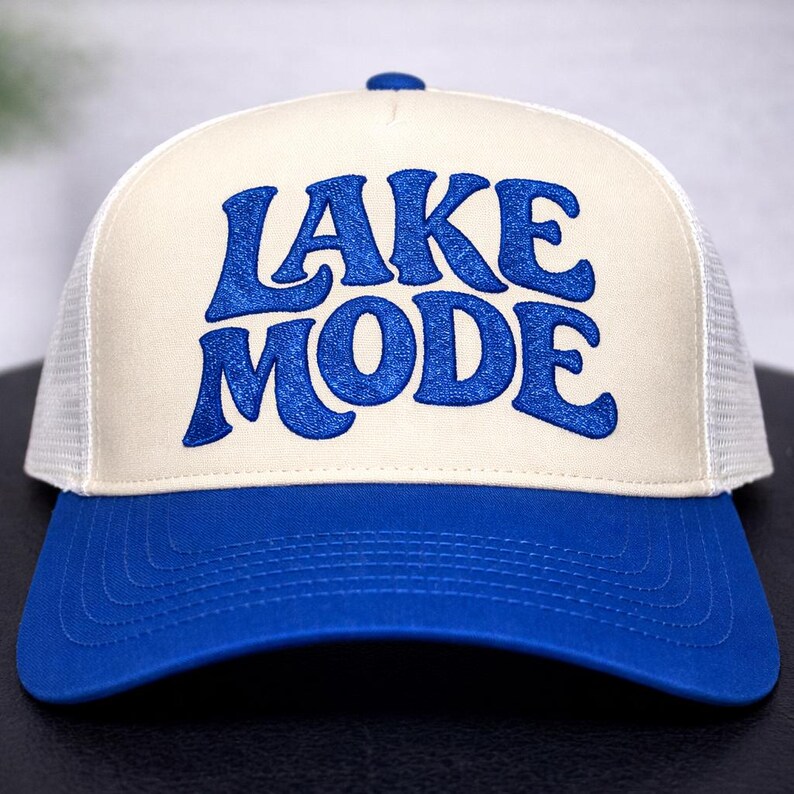 May include: A blue and cream-colored trucker hat with the words "LAKE MODE" embroidered in blue. The hat features a blue brim and top button, a cream-colored front panel, and a white mesh back.