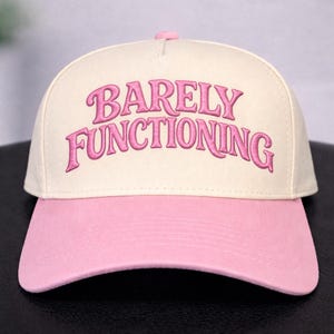 May include: A baseball cap with a cream-colored crown and a pink brim. The phrase "BARELY FUNCTIONING" is embroidered in pink script across the front of the hat. The hat has a curved brim and a pink button on top.