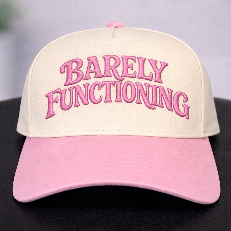 May include: A baseball cap with a cream-colored crown and a pink brim. The phrase "BARELY FUNCTIONING" is embroidered in pink script across the front of the hat. The hat has a curved brim and a pink button on top.