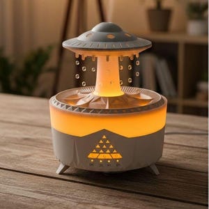 Puede incluir: Difusor de aceites esenciales gris y blanco con forma de OVNI y base naranja brillante. El difusor tiene un diseño único con luces y función de nebulización. Se encuentra sobre una superficie de madera, creando un ambiente cálido. Mide unos 20 cm de altura.