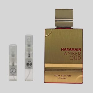 May include: A gold-colored perfume bottle with the text "Haramain Amber Oud Ruby Edition" and two small sample spray bottles. The large bottle has a rectangular shape with a gold cap and a red accent. The sample bottles are clear with white labels.