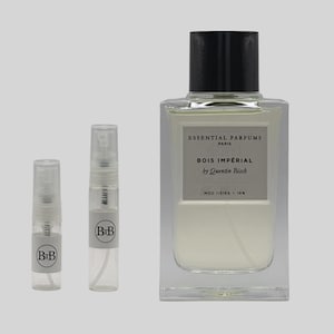 May include: A clear rectangular perfume bottle with a black cap, labelled "ESSENTIAL PARFUMS PARIS BOIS IMPÉRIAL by Quentin Bisch". Two small sample spray bottles with "BiB" labels are also present. The perfume has a light-coloured liquid.