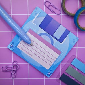 May include: A blue and purple floppy disk-shaped notepad with the text "GLITCHBERRY" and lined paper. A blue pen rests on the notepad, with purple paper clips and washi tape in the background. The notepad is on a purple tiled surface.