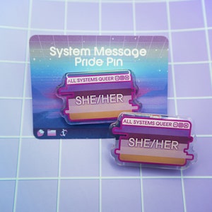 May include: Two clear acrylic pins with the text "SHE/HER" and "ALL SYSTEMS QUEER" in white. The pins have a purple, pink, and tan color scheme. The pins are on a backing card that reads "System Message Pride Pin".