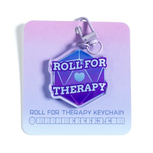 May include: Purple and blue gradient keychain with a dice shape and the text "Roll for Therapy" with a blue heart in the center. The keychain is attached to a silver metal ring.