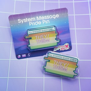 May include: A clear plastic pin with a green, yellow, and orange stripe design. The pin has the text "ALL SYSTEMS QUEER" and "THEY/THEM" in a white font.