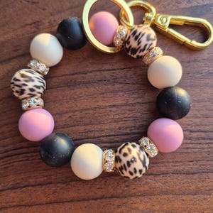 Peut inclure: Un bracelet porte-clés à perles avec des perles noires, blanches, roses et à imprimé léopard. Il est doté d'un fermoir et d'un anneau dorés. Les perles sont entrecoupées de petits espaceurs dorés scintillants.