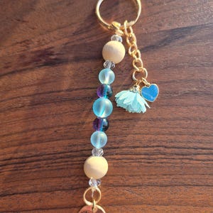 Peut inclure: Un porte-clés artisanal avec un anneau et une chaîne dorés. Il comprend des perles en bois, des perles bleues et violettes irisées et un petit charme en forme de cœur bleu. Un petit charme doré avec un motif floral est suspendu en bas.
