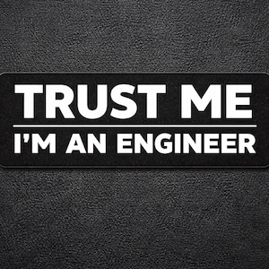 May include: A black rectangular sticker with the text "TRUST ME" above "I'M AN ENGINEER" in white, sans-serif font. The text is centered on the sticker, which is set against a textured black background.