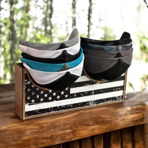 May include: A display of mesh trucker hats in various colors, including gray, white, black, and teal, stacked on a wooden box. The box is decorated with a distressed American flag design. The hats have a curved brim and a button on top.