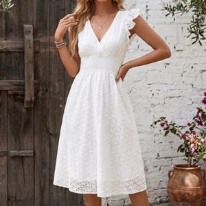 Peut inclure: Robe midi blanche en broderie anglaise avec un col en V, des manches à volants et une taille ajustée. La robe arrive au genou et a une jupe évasée. En arrière-plan, une porte en bois, un mur de briques blanches et un pot en terre cuite avec des fleurs.