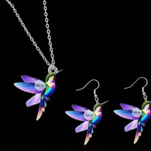 Peut inclure: Un ensemble de bijoux avec un motif de colibri. L'ensemble comprend un collier et deux boucles d'oreilles. Le colibri a des ailes et un corps violets, bleus, verts et jaunes. Les bijoux sont sur fond noir.