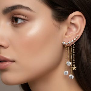 Peut inclure: Manchette d'oreille dorée ornée d'une rangée incurvée de pierres précieuses rondes et transparentes. Trois pierres précieuses rondes et transparentes et un petit charme en forme d'étoile pendent de la manchette, suspendus par de fines chaînes.