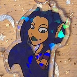 May include: A clear acrylic charm depicting a cartoon mermaid with blue hair and dark green lipstick. The charm has a rose gold-coloured keyring at the top. The mermaid's tail is a dark blue colour. The charm has a holographic effect.