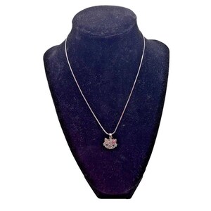 Puede incluir: Un collar plateado con un colgante con forma de cara de gato adornado con pequeños cristales brillantes. El collar se exhibe sobre un busto de terciopelo negro. El colgante mide aproximadamente 1,5 cm de ancho.