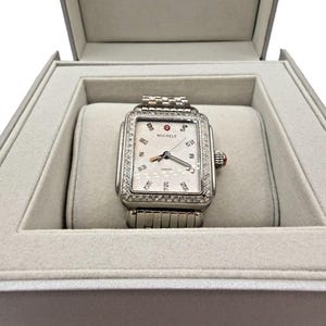 Puede incluir: Un reloj rectangular plateado con un bisel engastado con diamantes y una esfera blanca, presentado en una caja gris. El reloj tiene una correa plateada y un pequeño detalle rojo en el lateral. La marca "MICHELE" es visible.