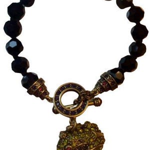 Puede incluir: Una pulsera con una hilera de cuentas negras facetadas conectadas a un cierre de barra circular con detalles morados. Un colgante con forma de rana, incrustado con pequeños brillantes verdes y rosas, cuelga del cierre metálico de la pieza.
