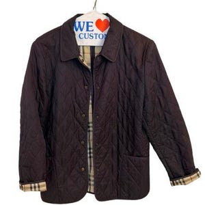 May include: A dark brown quilted jacket featuring a classic collar, snap button closures, and long sleeves with folded cuffs. The interior lining and the folded cuffs display a beige, black, and red plaid pattern. The jacket hangs on a white hanger.