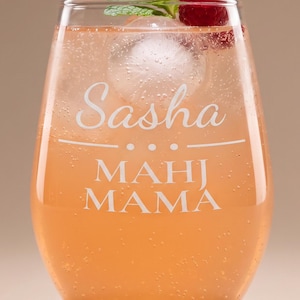 May include: A stemless glass filled with a light orange carbonated beverage, a round ice sphere, a fresh raspberry, and a sprig of mint. The glass is etched with the white text "Sasha" above a horizontal line and the words "MAHJ MAMA" below.
