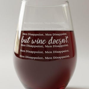 Può includere: Un bicchiere da vino senza stelo riempito di vino rosso. Il bicchiere presenta la frase "but wine doesn't" in corsivo, con le parole "Men Disappoint" ripetute sopra e sotto il testo principale. Il bicchiere è trasparente.