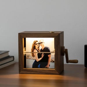 May include: A wooden photo cube with a hand crank, displaying a backlit photograph of a couple. The cube is made of dark wood and has a brass-colored button on top. The photo is visible through a clear panel.
