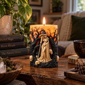 May include: A cylindrical orange candle featuring a relief sculpture of a figure in a wide-brimmed hat holding a staff, surrounded by black gnarled trees and small skulls. The candle is lit, casting a warm glow from the center of the wax structure.