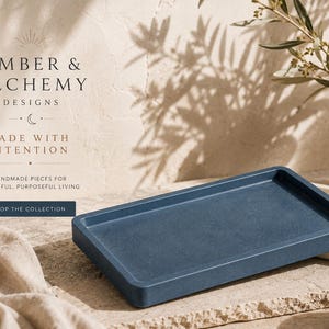May include: A rectangular dark blue tray with raised edges. The text on the image reads: EMBER & ALCHEMY DESIGNS, MADE WITH INTENTION, HANDMADE PIECES FOR BEAUTIFUL, PURPOSEFUL LIVING, SHOP THE COLLECTION. A second light beige tray is partially visible.
