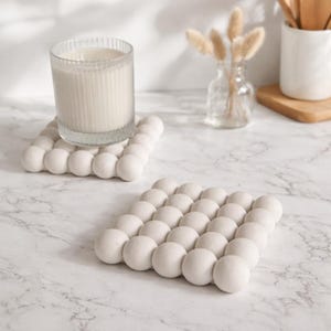 May include: Two off-white, square coasters with a bubble design. One coaster holds a clear glass candle holder with a white candle inside. The coasters are made of a solid material and are on a marble surface.