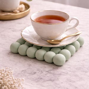 May include: A white teacup with a gold rim and a matching saucer, filled with tea, sits atop a light green sphere-shaped trivet. A gold spoon rests on the saucer. The scene is set on a light-colored surface.