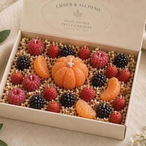 May include: A rectangular gift box containing assorted fruit-shaped candles, including a central orange pumpkin, segments of mandarin, red raspberries, and black blackberries. The lid features the text Ember & Alchemy Hand-Poured Fruit Candles Natural Soy Wax.