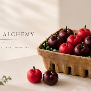May include: A brown fiber basket filled with red and dark purple apple-shaped candles, accompanied by two loose apple candles and three small blue berry-shaped candles. The text Ember & Alchemy Handcrafted Candles & Curiosities is visible.