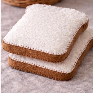 Puede incluir: Dos rebanadas de pan blanco con corteza marrón, apiladas una encima de la otra. El pan tiene un aspecto texturizado y esponjoso y está cosido alrededor de los bordes con hilo blanco. Una cesta de mimbre está en el fondo.