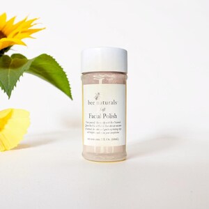 May include: A white jar with a pink label that says "bee naturals Soft Facial Polish". The jar is sitting on a white surface. The text on the label says "Treat yourself daily with Bee Naturals Queen Bee Facial Polish. Our all-natural formula is a gentle & grainy exfoliating clay that brightens and evens your complexion. Net contents 2.1 Oz. (60ml)"