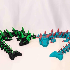 May include: Two articulated dragon figures. One is black with green accents, the other is teal with red accents. Each dragon has a head, body, and tail, and is designed with a gaming controller at the end of the tail.