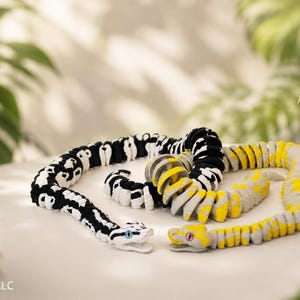 May include: Three toy snakes on a white surface, with a backdrop of foliage. One is black and white, another yellow and grey, and the third is black and yellow. The snakes have detailed heads and are coiled.