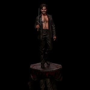 May include: A figurine of a man in a black leather shirt and pants, holding a sword. The figure is standing on a stone platform with red accents, against a black background. The man is smiling.