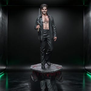May include: A statue of a man in a black leather shirt and pants, holding a sword. The statue stands on a stone platform with red accents. The man has dark hair and is smiling. The background is dark with green lighting.